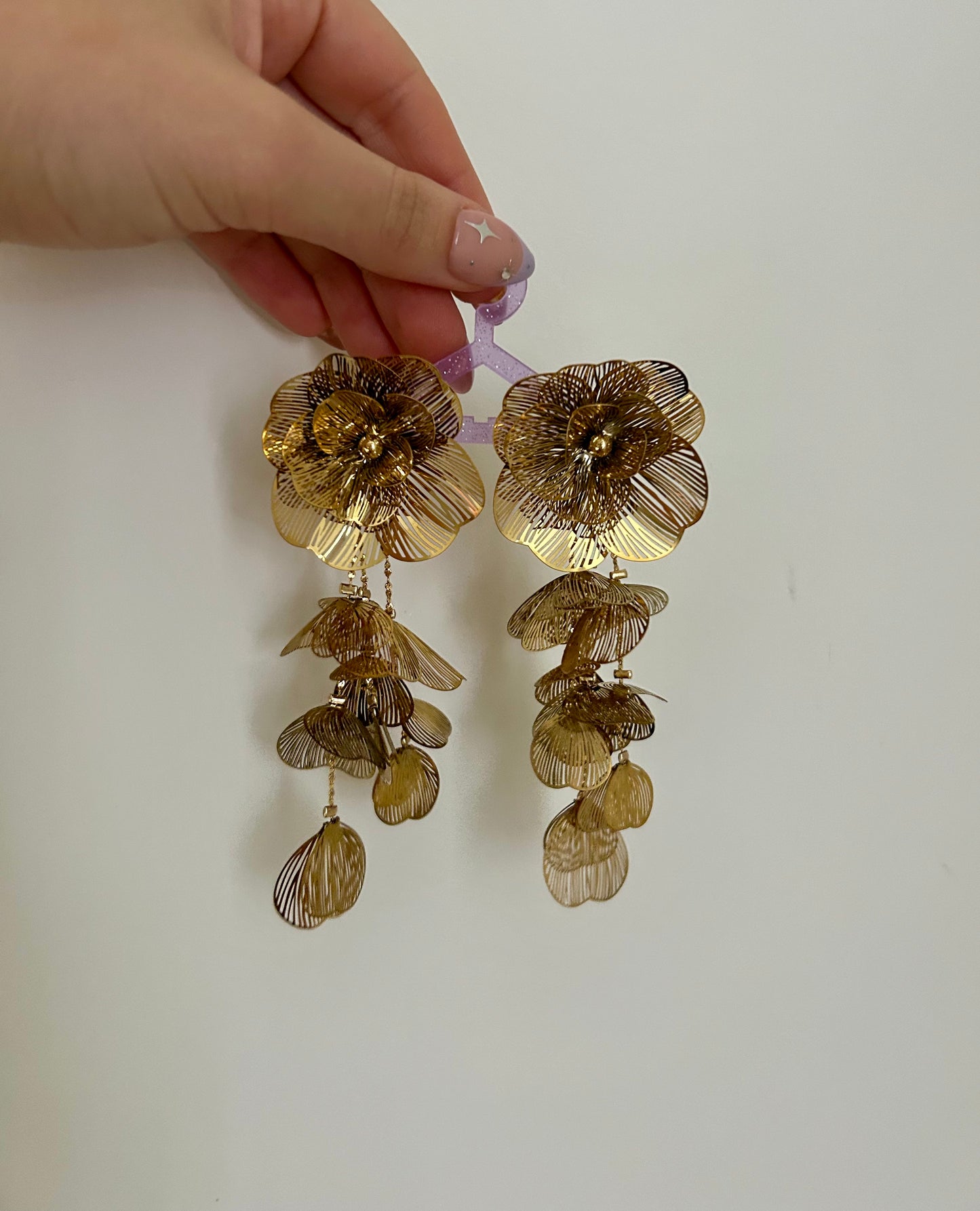 Gold floral earrings held by a hand against a plain background