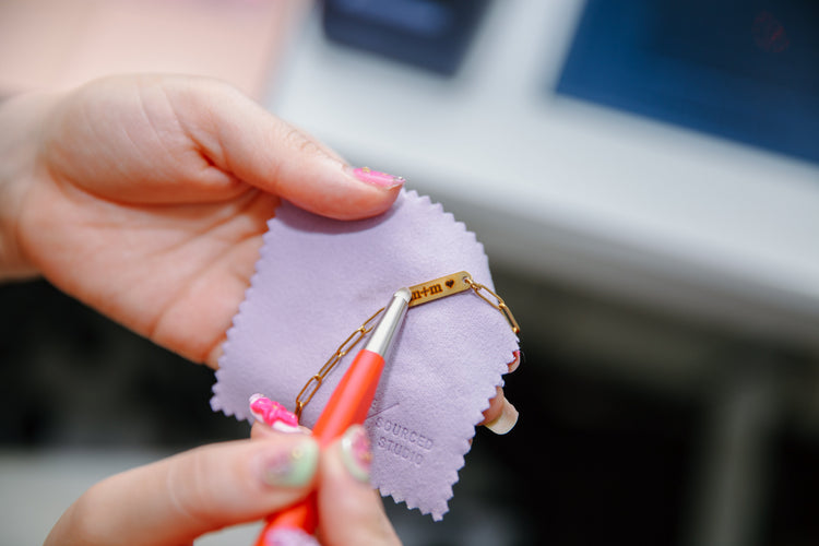 A close-up of a person's hands holding a small gold chain tag or keychain on a light purple cleaning cloth, using a small orange tool to clean or polish it.