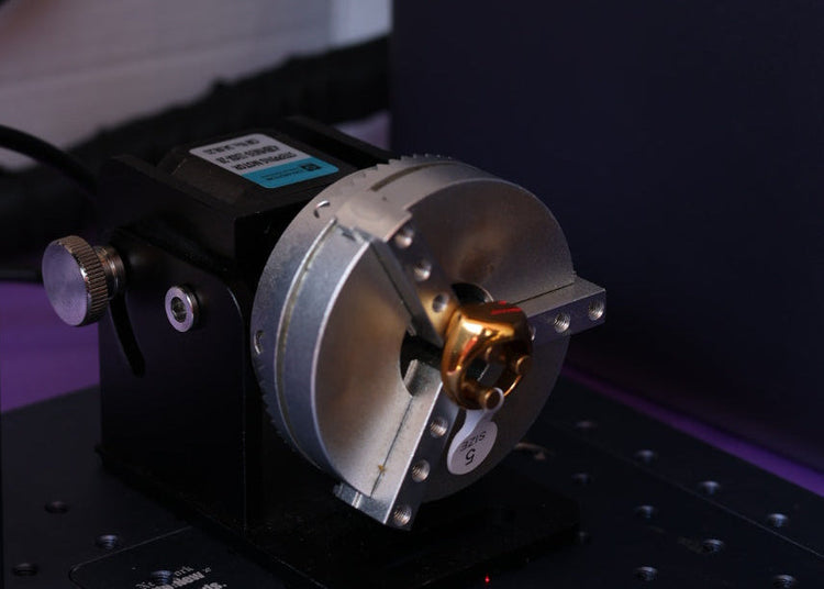 A close-up of a laser marking head mounted on a black metal bedplate, with a small gold-colored workpiece held in the center. The background shows a dark machine body and some cables.