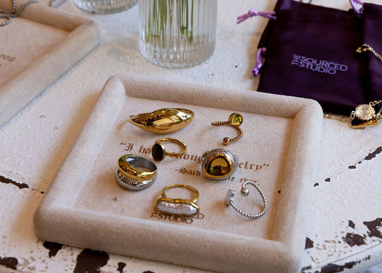 A styled jewelry display on a distressed white table, featuring gold and silver rings on a cream-colored tray, with a dark purple velvet jewelry pouch branded 'LUXE JEWELRY' and two glass vases with flowers in the background.