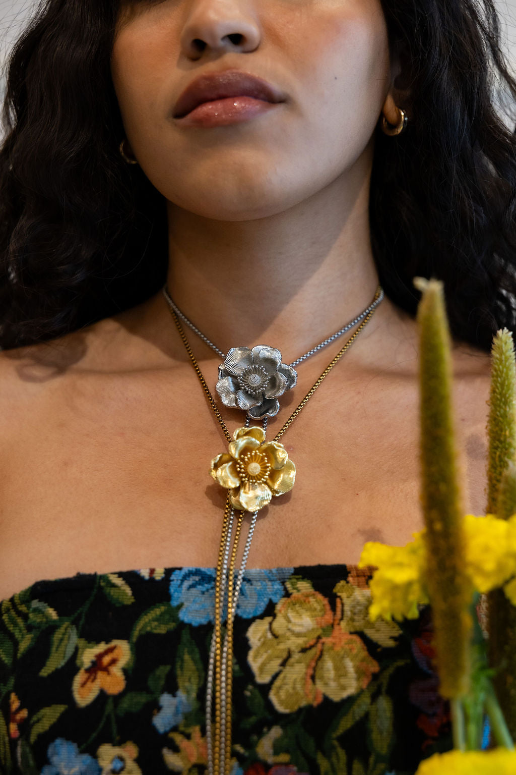 A close-up of a model wearing a layered necklace with two flower charms, one silver and one gold, over a floral strapless top. Yellow flowers are visible in the foreground.
