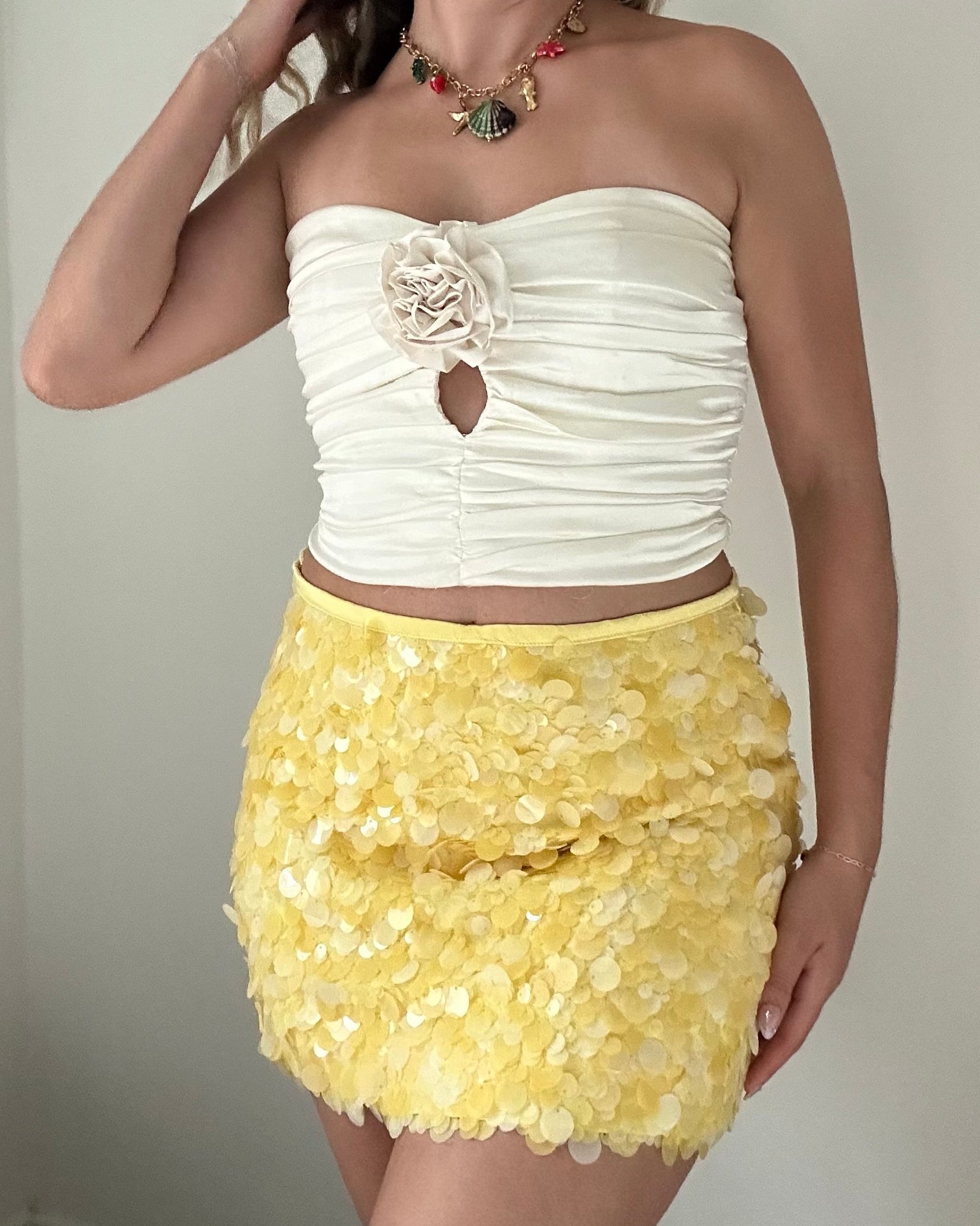 Woman wearing a white strapless top with floral detail and a yellow sequin skirt against a neutral background.