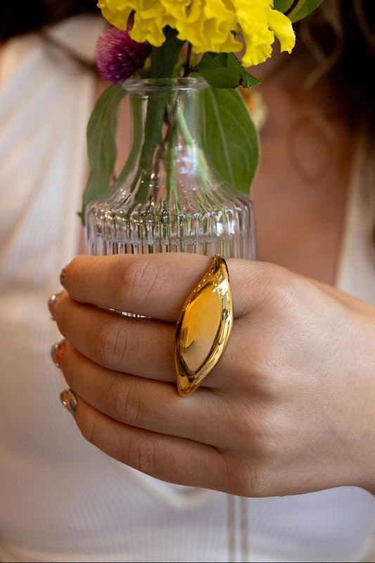 A close-up lifestyle shot of a woman holding a small clear glass vase with yellow flowers. The woman is wearing a large, prominent gold ring with a marquise-shaped yellow stone on her finger.