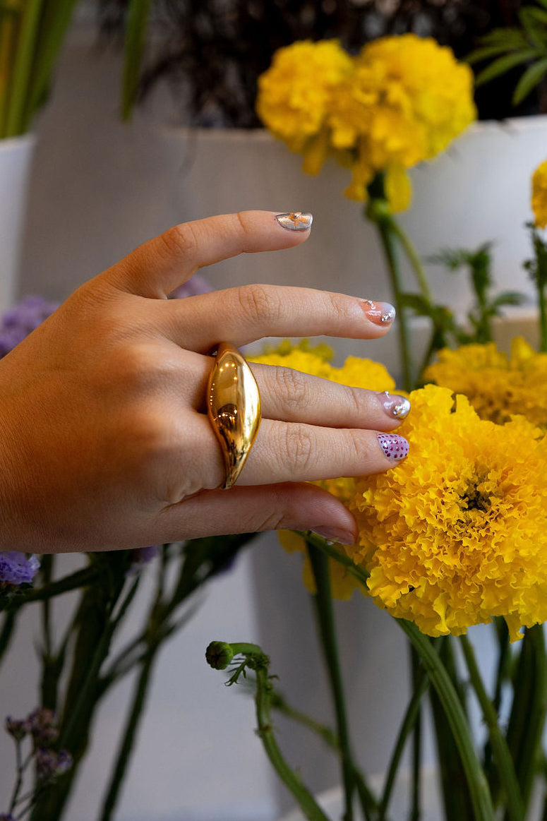 A close-up of a hand wearing a large, polished gold ring, gently touching bright yellow marigold flowers in white pots.
