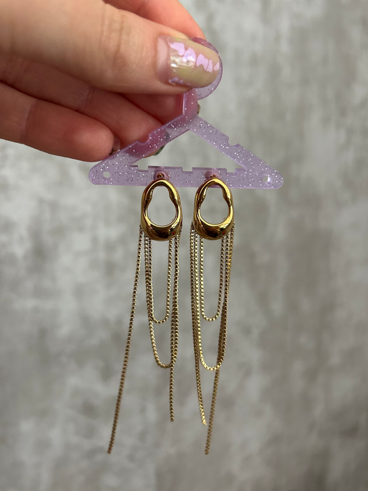 Gold chain earrings on a clear plastic stand held by a hand with glittery nail polish against a neutral background.