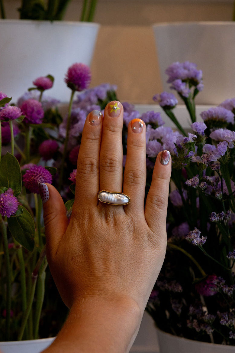 A close-up of a hand wearing a silver ring with a white pearl-like center, resting near purple flowers in white pots.