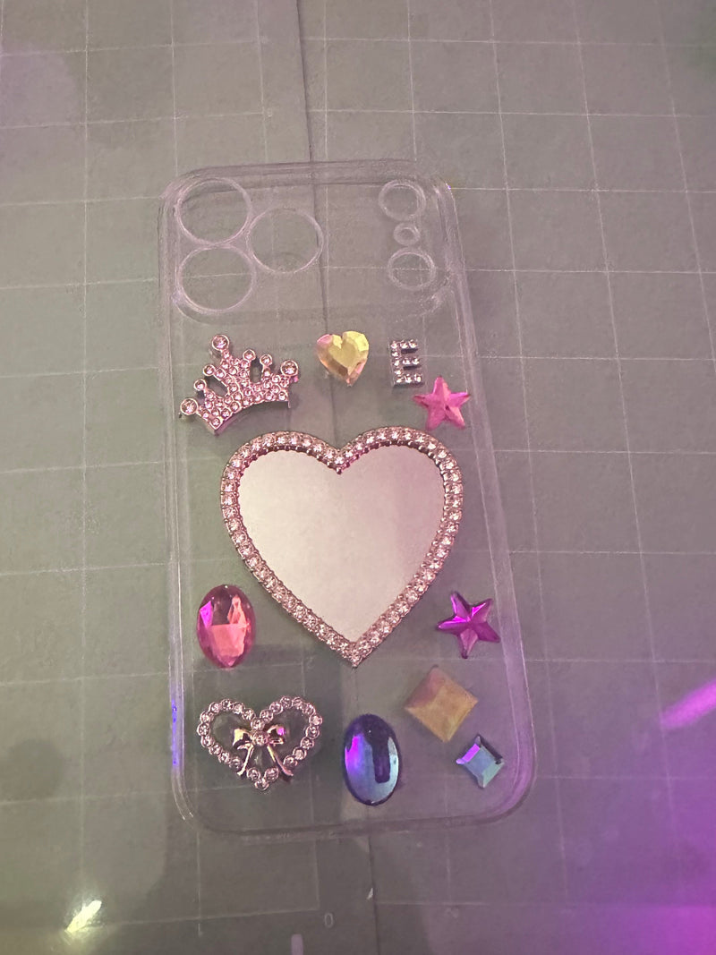 A clear phone case decorated with a large rhinestone-embellished heart, smaller hearts, stars, and gem-like accents, shown on a gray gridded cutting mat with purple lighting.