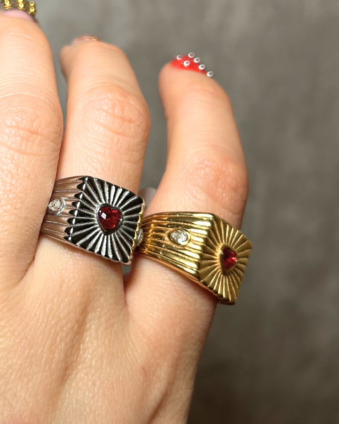 Two rings with heart designs and red stones on a finger against a neutral background