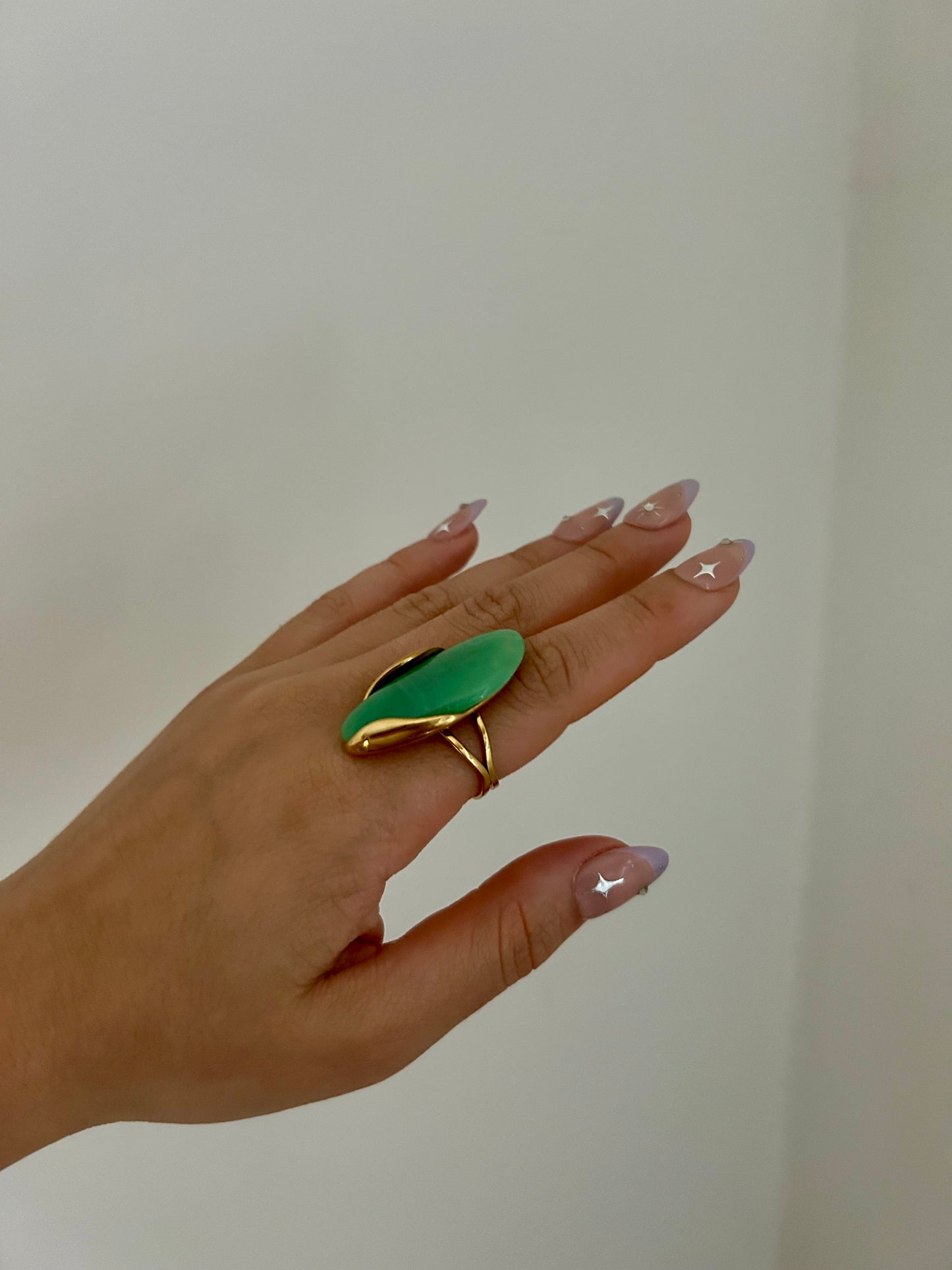 Hand wearing a green leaf-shaped ring with gold border on a plain background
