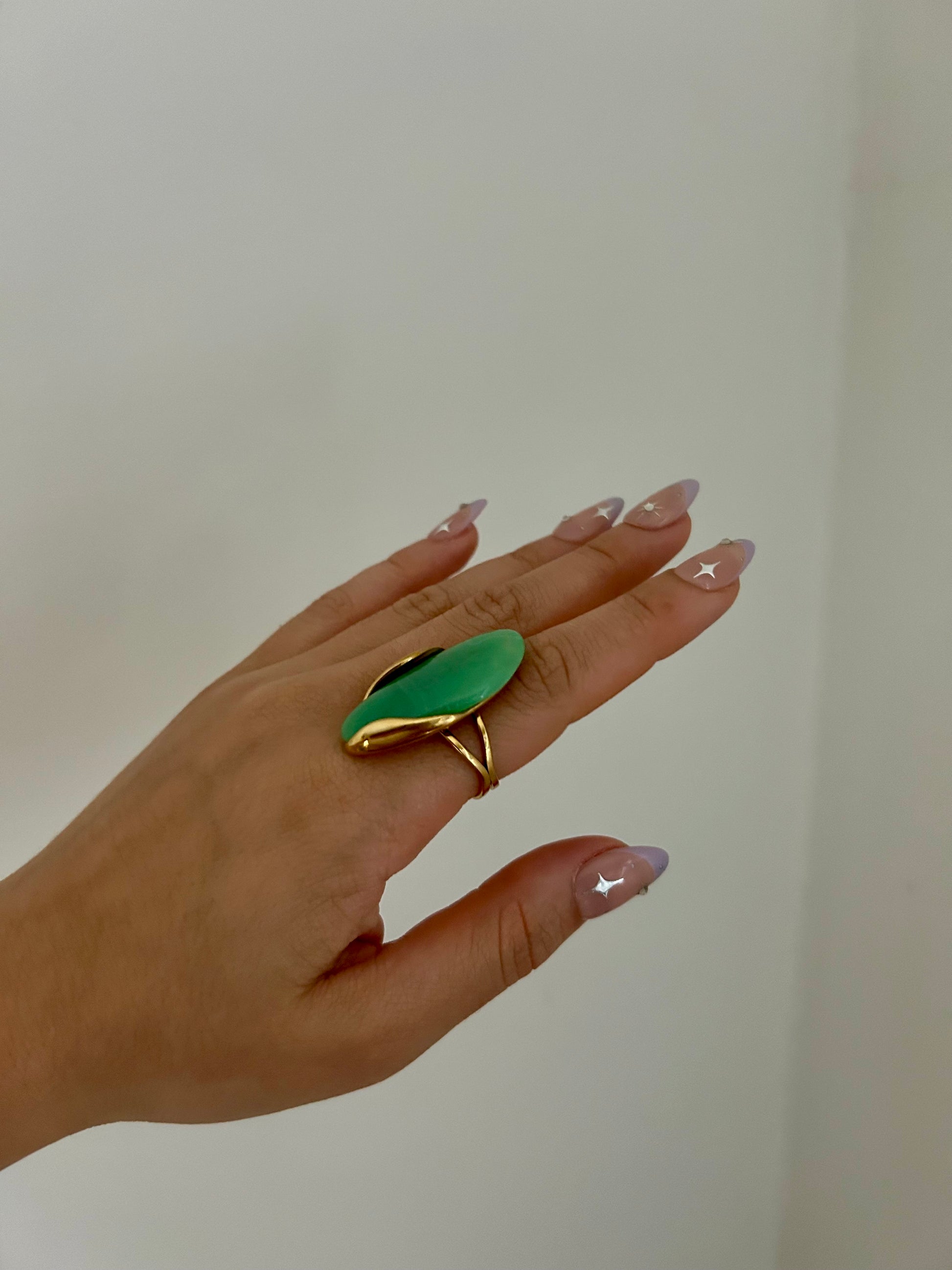 Hand wearing a green leaf-shaped ring with gold border on a plain background