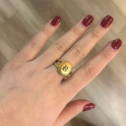 Hand wearing a gold ring with a logo, against a neutral background
