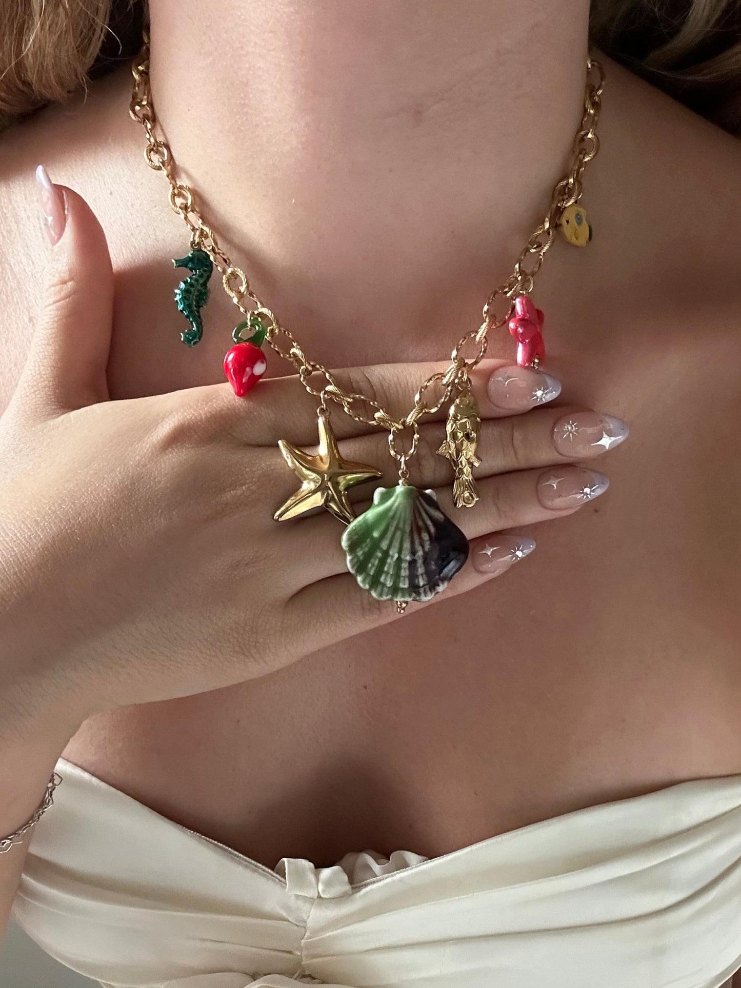 Close-up of a person wearing a gold bracelet and ring with colorful charms, wearing a white dress.