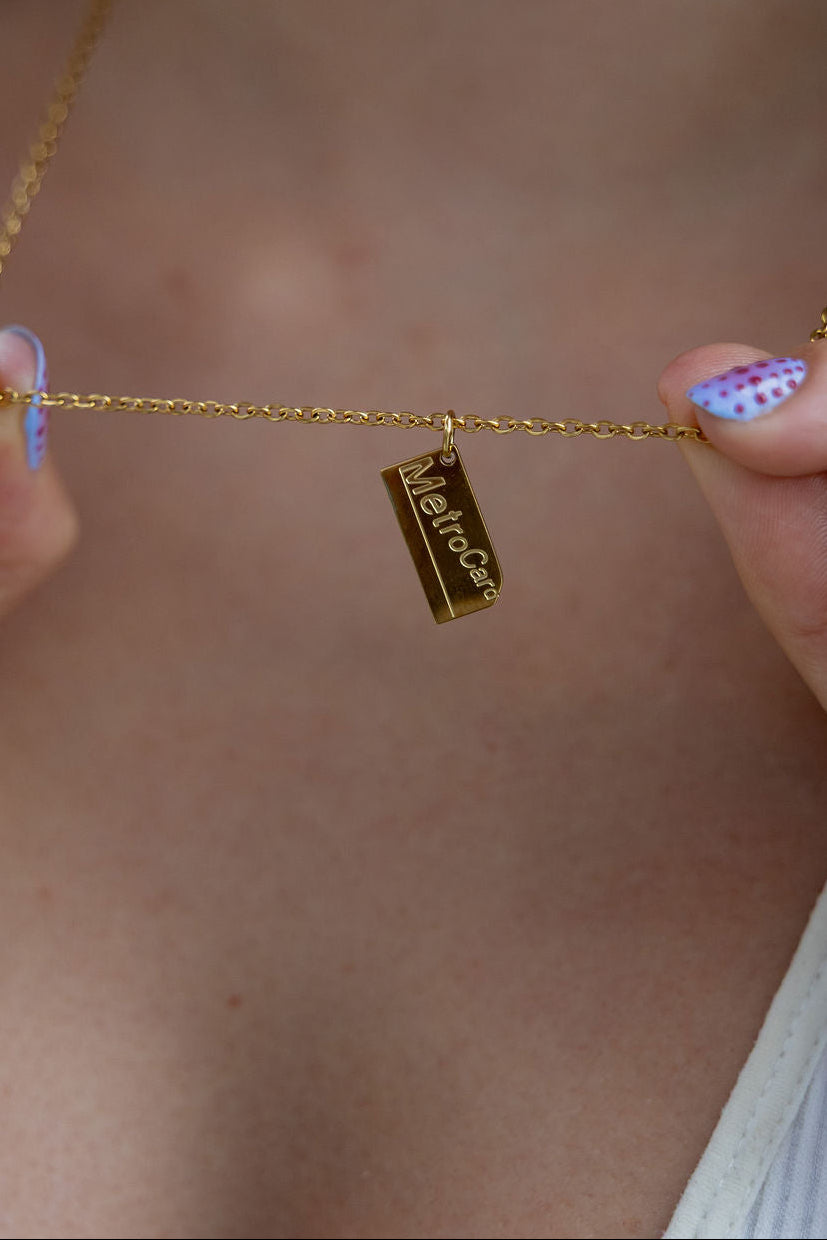 A close-up lifestyle shot of a person holding a delicate gold chain necklace with a small rectangular gold pendant engraved with the brand name 'MetroCard'.