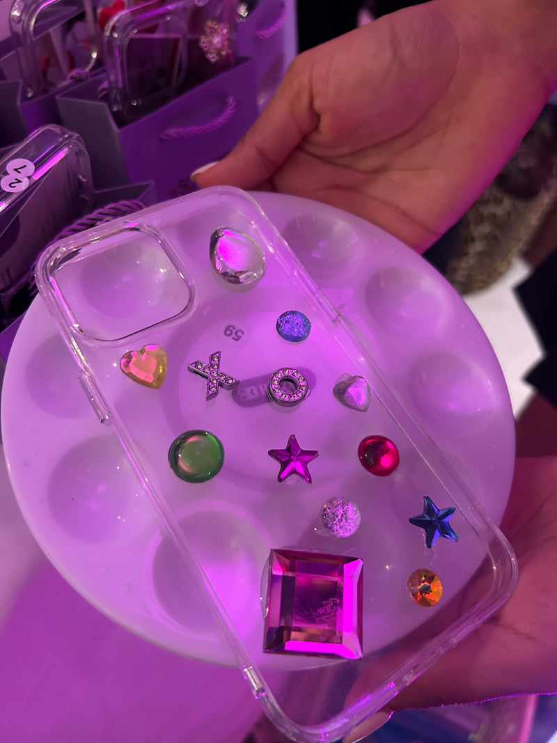 A clear phone case decorated with multiple colorful rhinestones and charms, held over a white dual-pigment paint palette under pink and purple lighting.