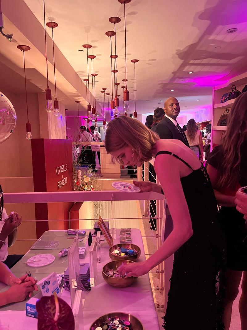 A woman in a black sleeveless dress leans over a table at what appears to be a boutique or event space, surrounded by other shoppers and hanging pendant lights under warm pink ambient lighting.