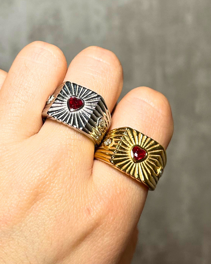 Two rings with sunburst designs and red stones on a finger against a neutral background
