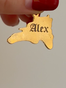 Gold state-shaped pendant with 'Alex' engraved, held by a hand.