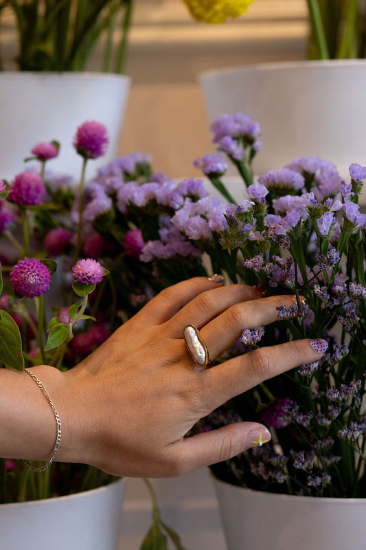 A close-up of a person's hand wearing a chunky silver ring, gently touching purple flowers in a white pot.