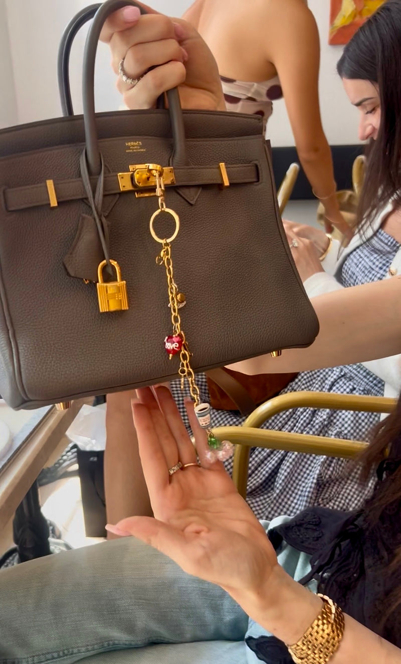 Brown handbag with gold hardware and keychain held by a person, with another person in the background.