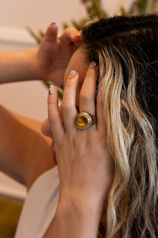 A close-up lifestyle shot of a person wearing a large silver ring with a round yellow stone, touching their hair with both hands.